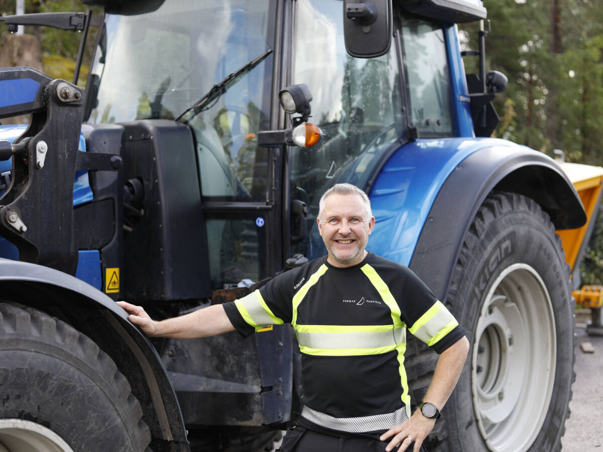 Maarakennusalan ammatti- ja erikoisammattitutkinnon suorittanut katutyönjohtaja seisoo iloisesti hymyillen kaupungin varikolla sinisen traktorin edessä.